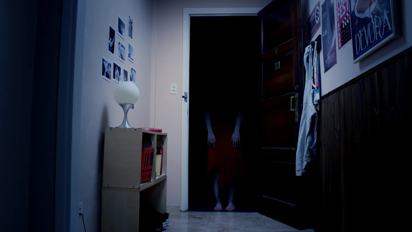 Fotograma de Tenebris Somnia mostrando una figura espectral en un pasillo oscuro frente a una puerta abierta.